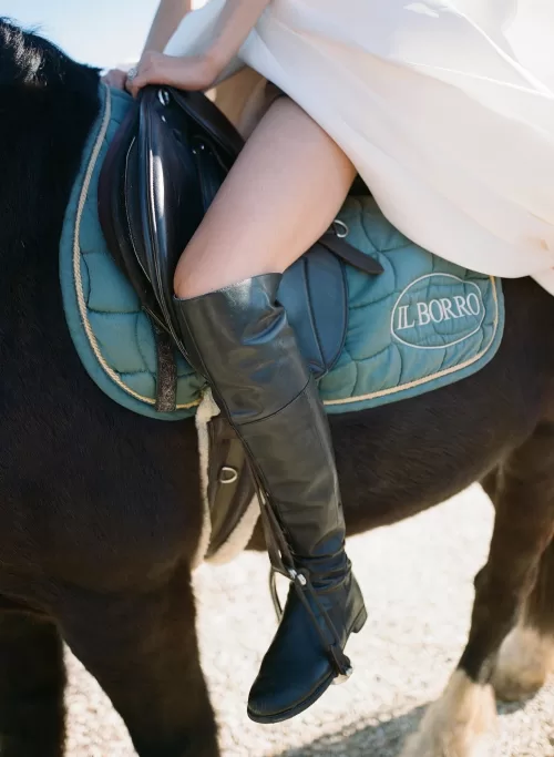 Bride in chic riding boots on horseback, the Il Borro saddle pad visible beneath soft sunlight.