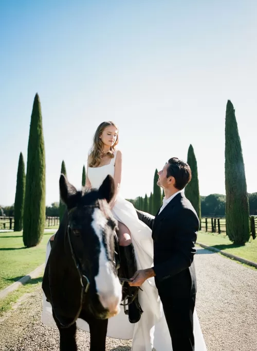 Groom helping bride seated on a black horse along Il Borro’s sunlit cypress avenue, evoking Tuscan romance.