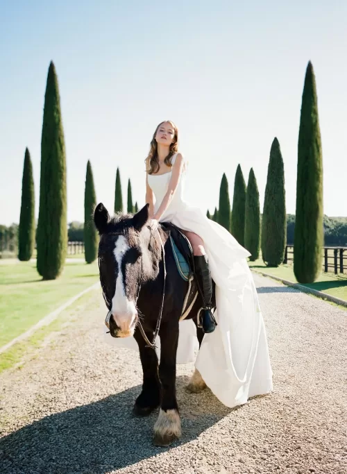 Bride sitting gracefully on a dark horse beneath Il Borro’s tall cypress trees, her gown flowing in the breeze.