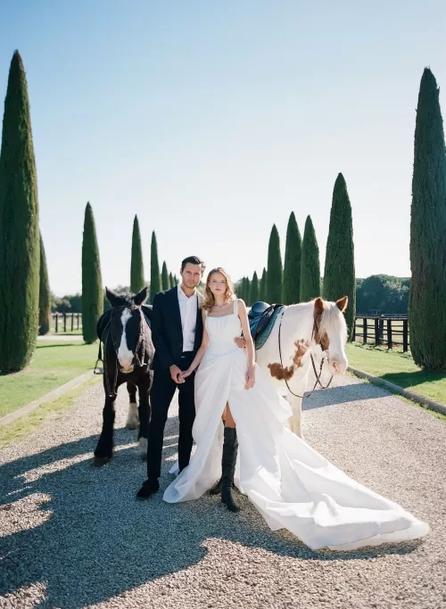 Bride and groom stand hand in hand with two horses on Il Borro’s gravel avenue framed by cypress trees.