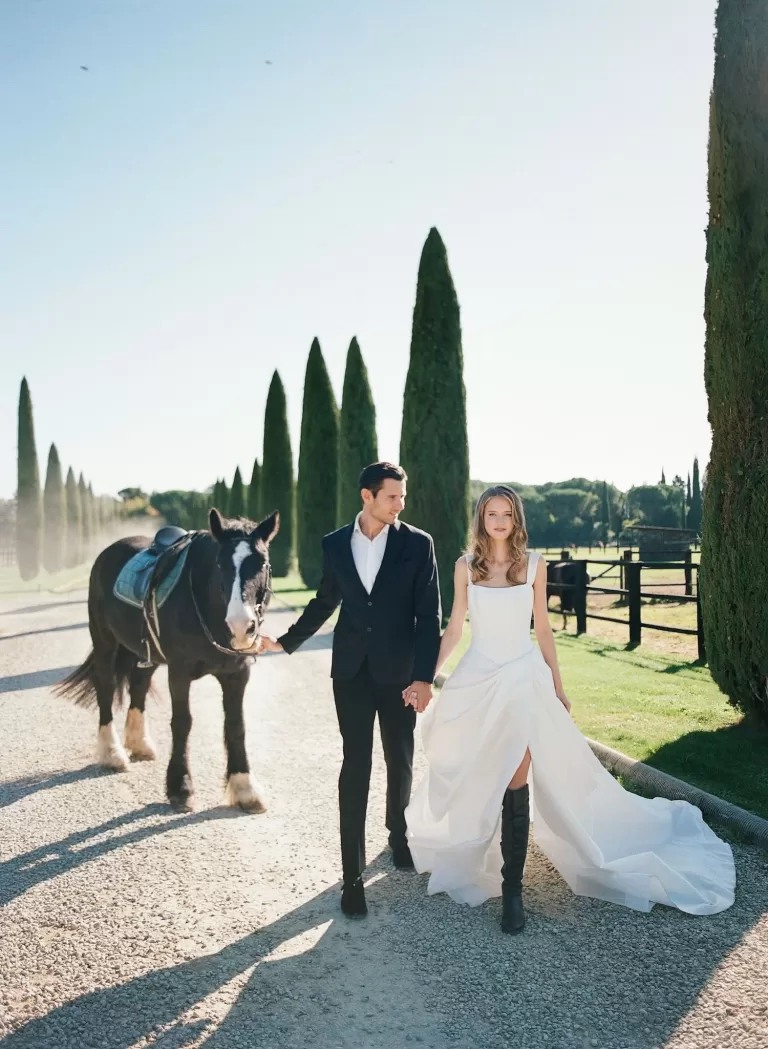 Bride and groom hand in hand leading a horse down Il Borro’s tree-lined gravel path in soft sunlight.
