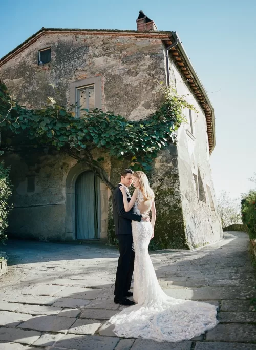 Bride and groom embrace beside a rustic Tuscan villa at Il Borro, sunlight highlighting her lace gown.