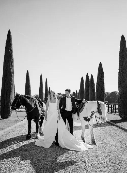 Bride and groom walk with two horses down the elegant cypress drive at Il Borro under clear Tuscan skies.