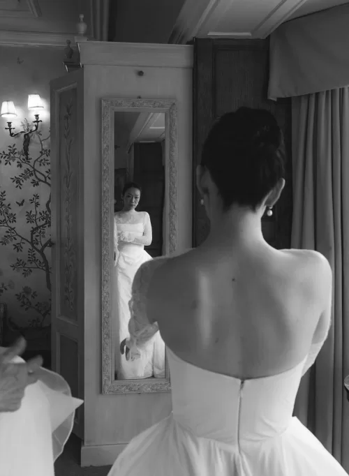 The bride admires her gown in a vintage mirror at Manor House, surrounded by elegant wallpaper and soft light.