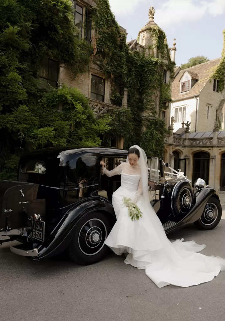 The bride steps gracefully from a classic black car at Manor House, holding her bouquet and veil.