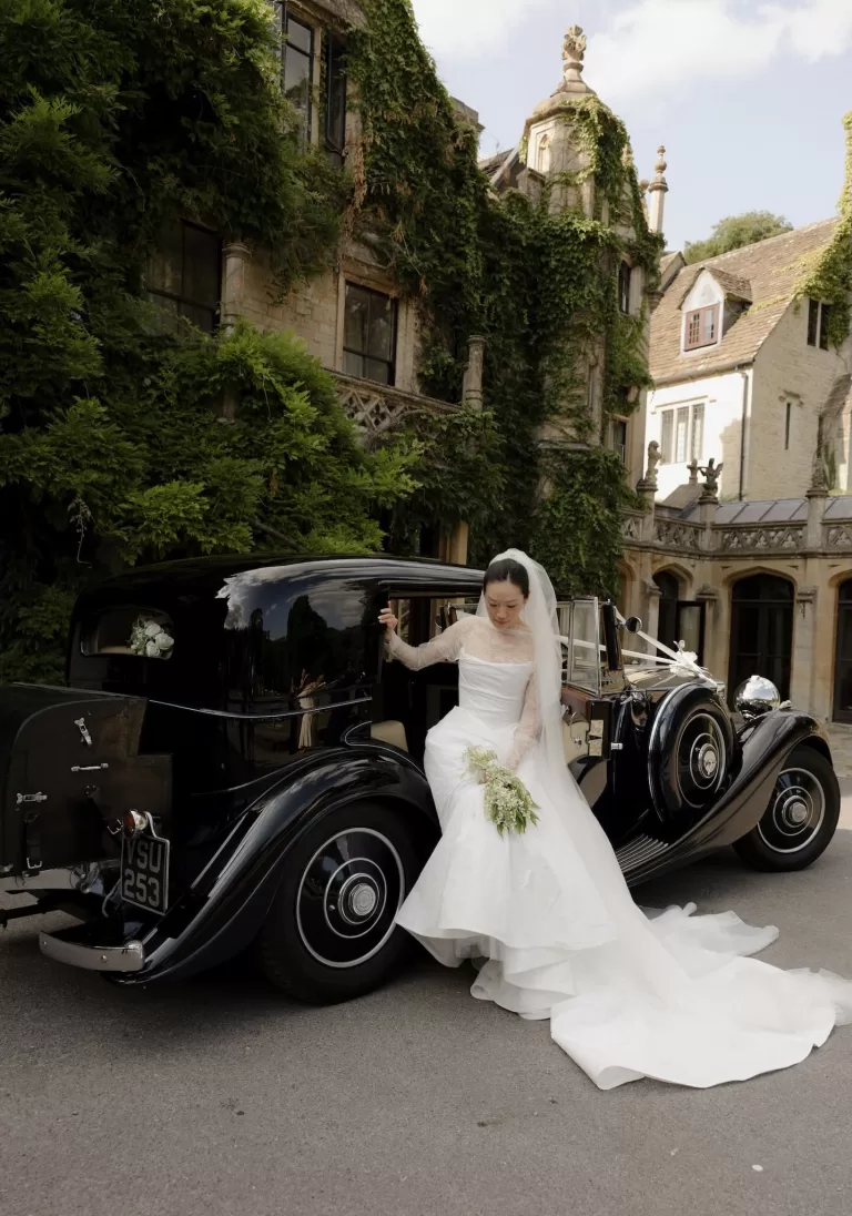 The bride steps gracefully from a classic black car at Manor House, holding her bouquet and veil.
