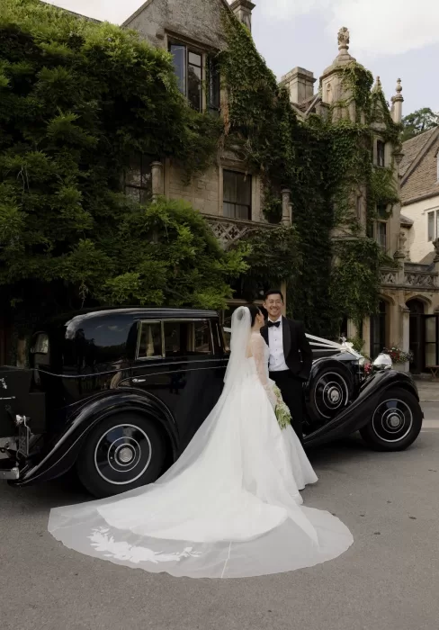 Bride and groom pose beside a vintage black car in front of the ivy-covered Manor House.