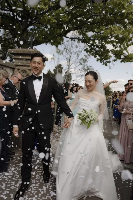 Newlyweds walk through a shower of white confetti at Manor House, smiling as guests celebrate their union.