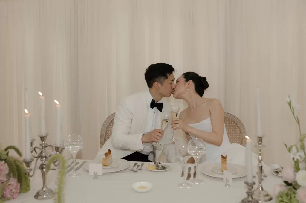 The couple toast with champagne and share a kiss at their softly lit Manor House reception dinner.