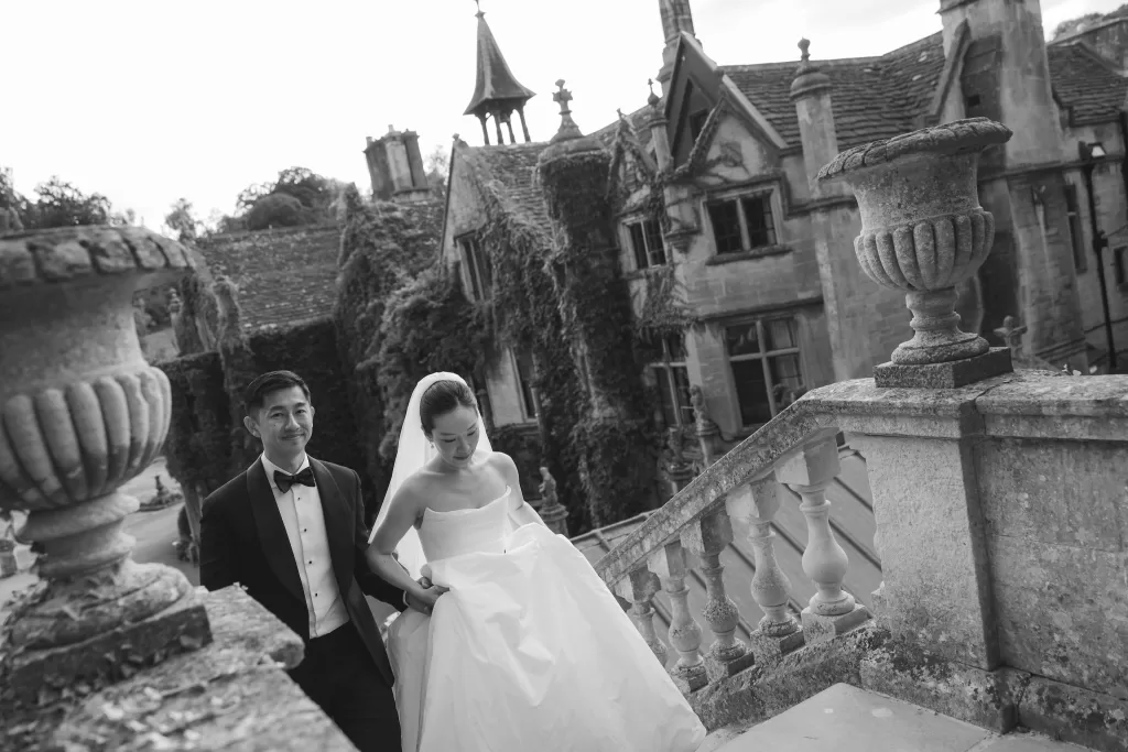 The couple walk up the grand stone staircase of Manor House, framed by ivy-covered walls in a timeless black and white moment.