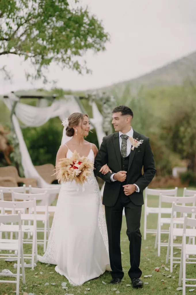 Bride and groom walking arm in arm through the ceremony garden at Masia Can Ramonet, smiling at each other after the vows.