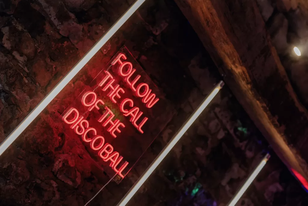 A glowing neon sign reading “Follow the Call of the Discoball” adds playful charm to the stone walls at This Must Be The Place.