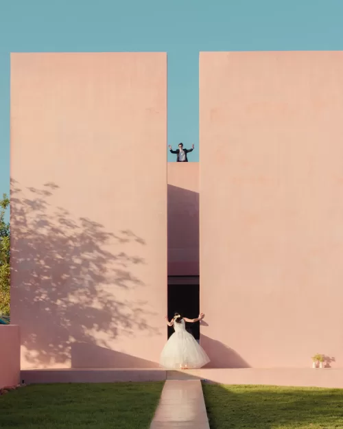 Bride in gown and groom above her framed by Neuendorf House’s striking pink walls and clear sky.