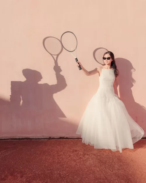 Bride in tulle gown poses with a tennis racket against the pink walls of Neuendorf House.