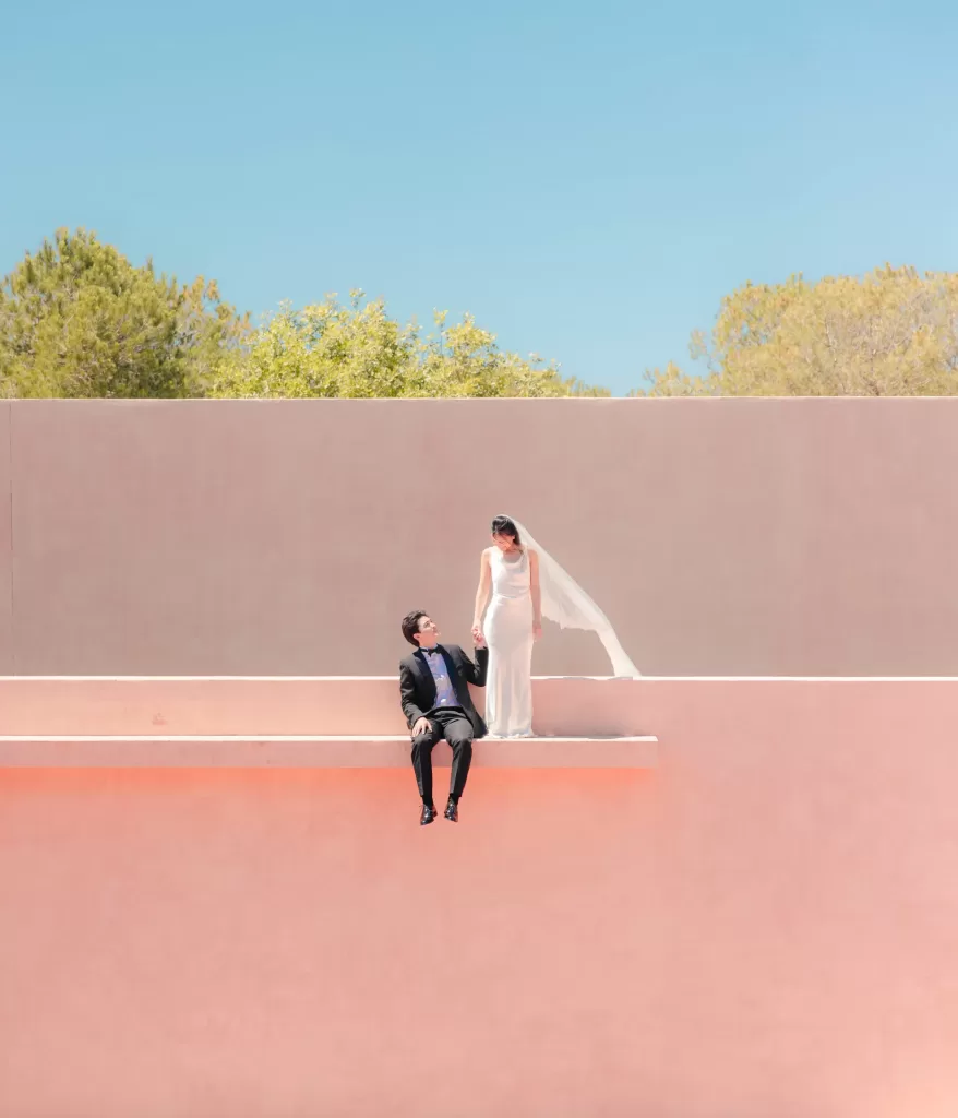 Bride and groom pose on the pink walls of Neuendorf House under a clear blue sky.