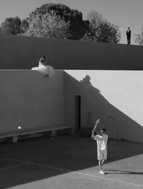 Groom plays tennis in sunlight while the bride sits above the court at Neuendorf House.