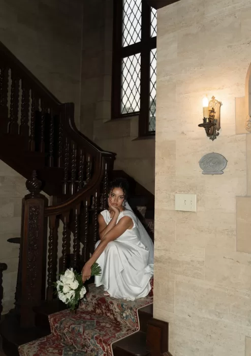 Bride in satin gown sits gracefully on the carved wooden staircase at The Plummer House.