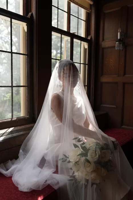 Bride sits by the window with her veil softly draped over her face, holding white roses at The Plummer House.