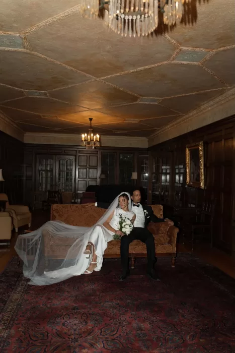 Bride and groom relax together on a vintage sofa under chandeliers in The Plummer House lounge.