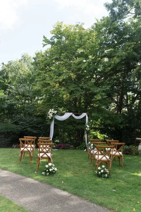 Elegant outdoor ceremony setup with white draped arch and wooden chairs in The Plummer House garden.