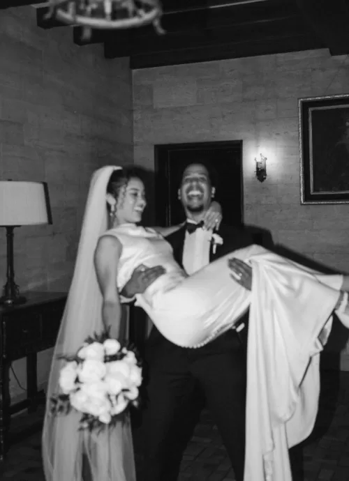 Groom joyfully carries his bride through the hallway of The Plummer House in a candid black and white shot.