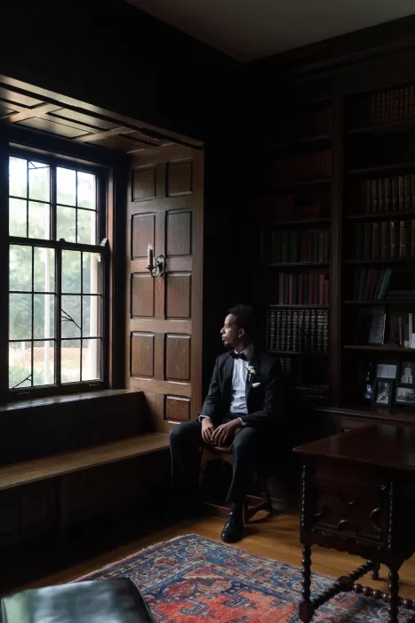 Groom sits by the window in the wood-paneled library, looking out at the grounds of The Plummer House.
