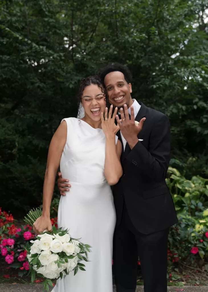 Newlyweds laugh and show their wedding rings while standing in the gardens of The Plummer House.