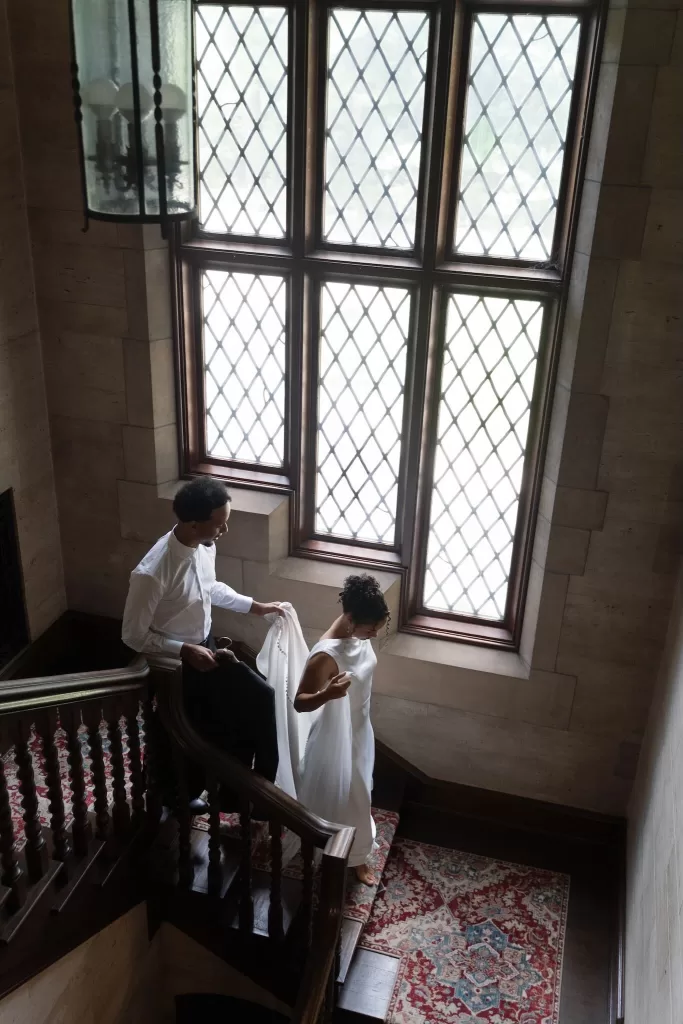 Groom gently helps the bride down the staircase filled with natural light at The Plummer House.