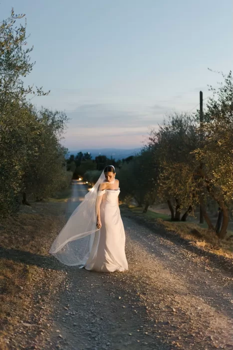 Bride standing on a country lane lined with olive trees at twilight at Quercia al Poggio.