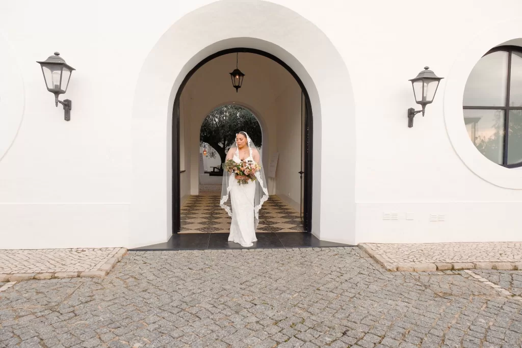 Bride walks through the elegant white arches of Quinta Dos Santos holding a romantic floral bouquet.