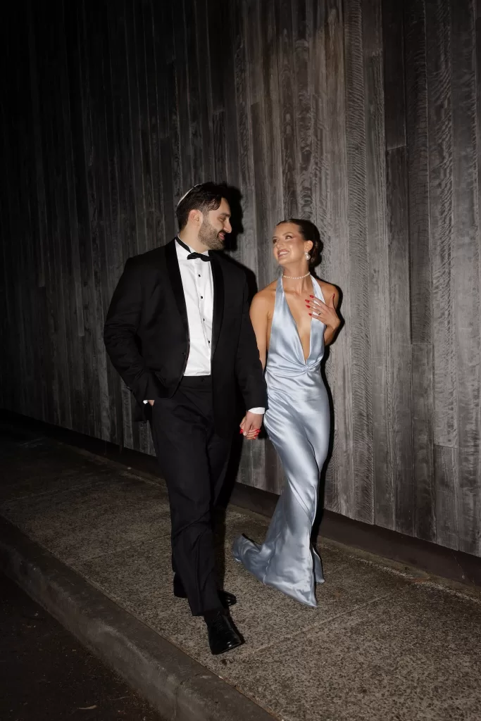 Bride in satin gown walking hand in hand with groom in tuxedo outside Sergeants Mess.