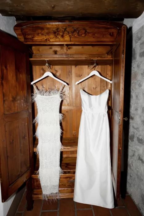 Two wedding dresses hanging in a rustic wooden wardrobe at This Must Be The Place, one classic and one feathered.