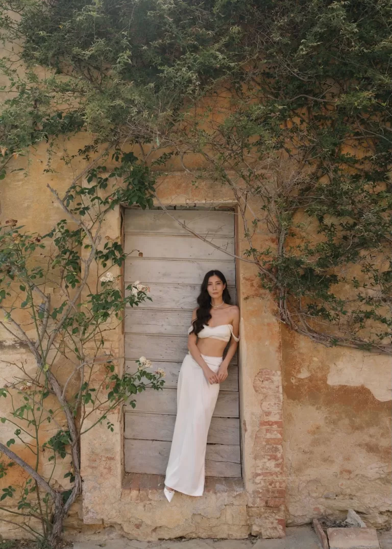 Bride in a two-piece ivory outfit leans against a rustic terracotta wall covered in greenery at Villa Catignano.