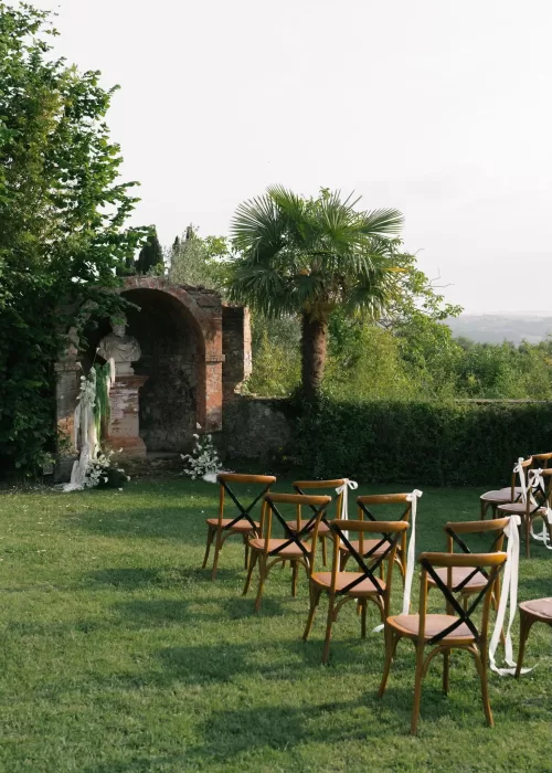 Outdoor wedding ceremony setup at Villa Catignano with wooden chairs lined on the lawn facing a stone arch and greenery.