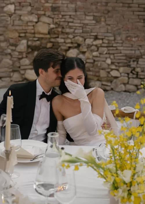 Bride laughs as the groom whispers in her ear during dinner at Villa Catignano, surrounded by yellow flowers and candlelight.