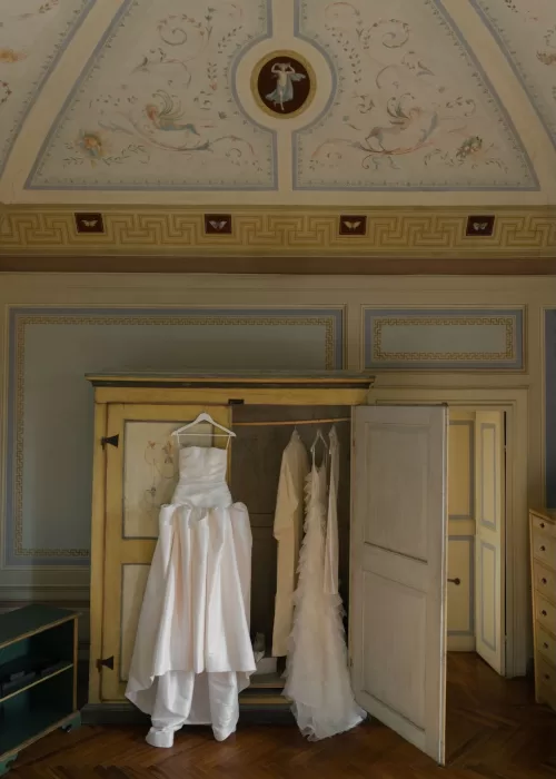 Wedding gowns hang elegantly in a painted armoire beneath an ornate ceiling at Villa Catignano.