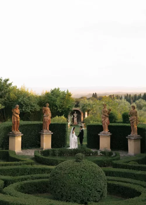 Bride and groom walk hand in hand between stone statues in the manicured gardens of Villa Catignano at sunset.