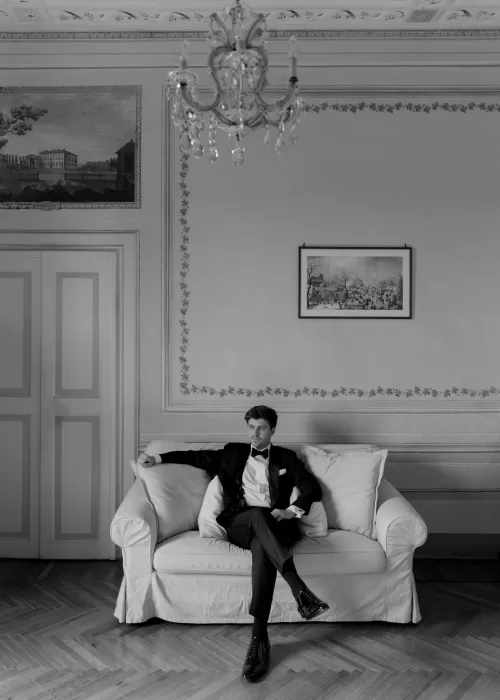 Black and white portrait of the groom in a tuxedo sitting on a cream sofa beneath a chandelier at Villa Catignano.
