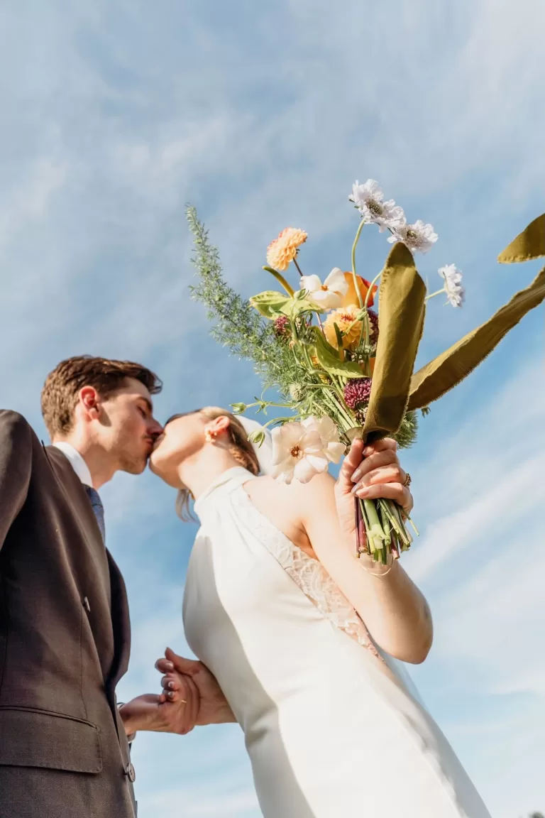 The newlyweds share a kiss under a clear blue sky at This Must Be The Place, with a bouquet of colorful flowers in hand.