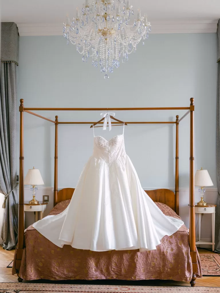 Elegant bridal gown hanging from a four-poster bed beneath a crystal chandelier inside a Château Bouffémont suite.