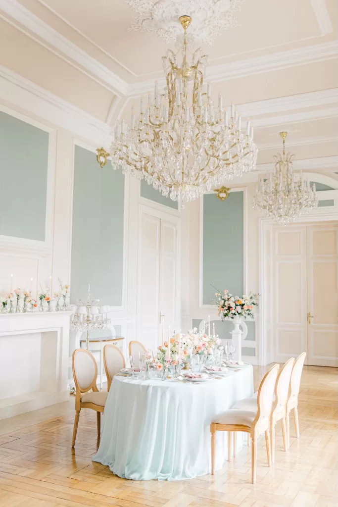 Round pastel reception table set beneath ornate chandeliers in Château Bouffémont’s refined salon.