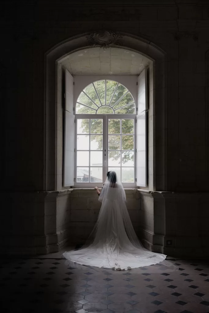 Bride in flowing veil standing before a tall arched window inside Château de Jalesnes, softly lit by daylight.