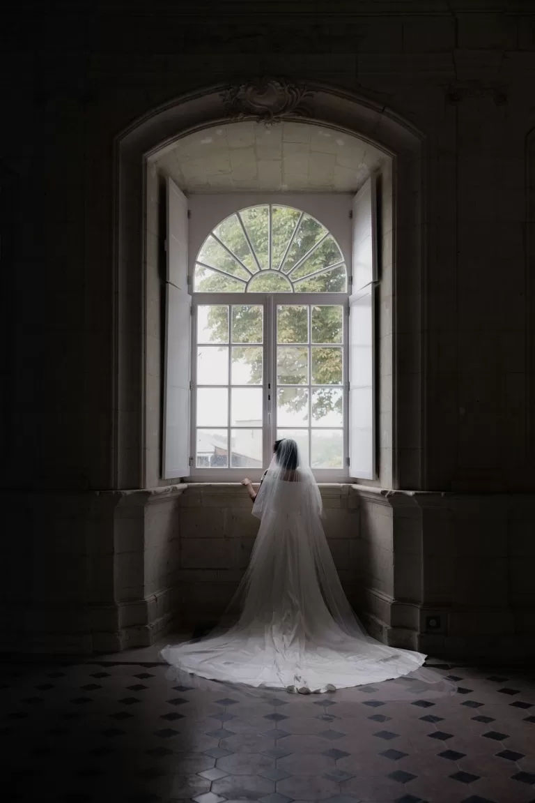 Bride in flowing veil standing before a tall arched window inside Château de Jalesnes, softly lit by daylight.
