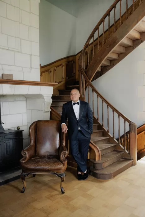 Groom in a black tuxedo posing beside a leather armchair and wooden staircase at Château de Jalesnes.