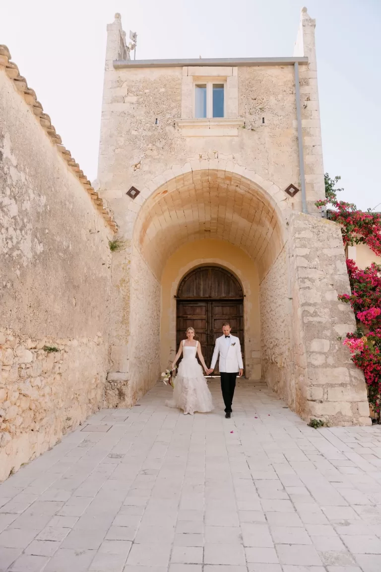 Bride and groom walking hand in hand through the grand stone archway of Dimora Delle Balze.