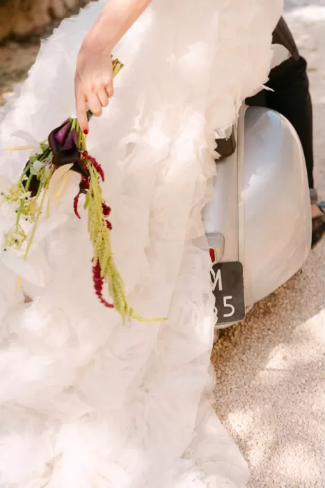 Bride’s hand holding a cascading floral bouquet while sitting on a vintage Vespa at Dimora Delle Balze.