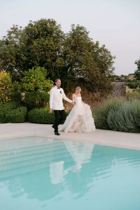 Bride and groom walking joyfully by the turquoise pool surrounded by gardens at Dimora Delle Balze.