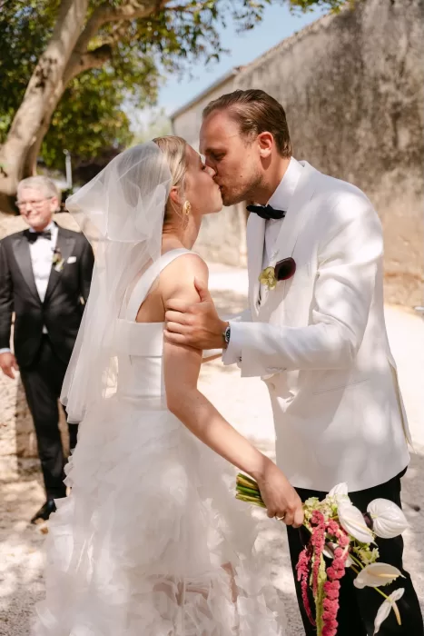 Newlyweds share a romantic kiss after the ceremony at Dimora Delle Balze, sunlight filtering through trees.