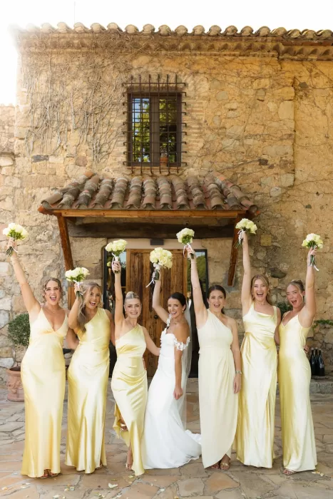 Bride celebrating with her bridesmaids in pale yellow gowns outside the rustic stone building at Mas del Sord.