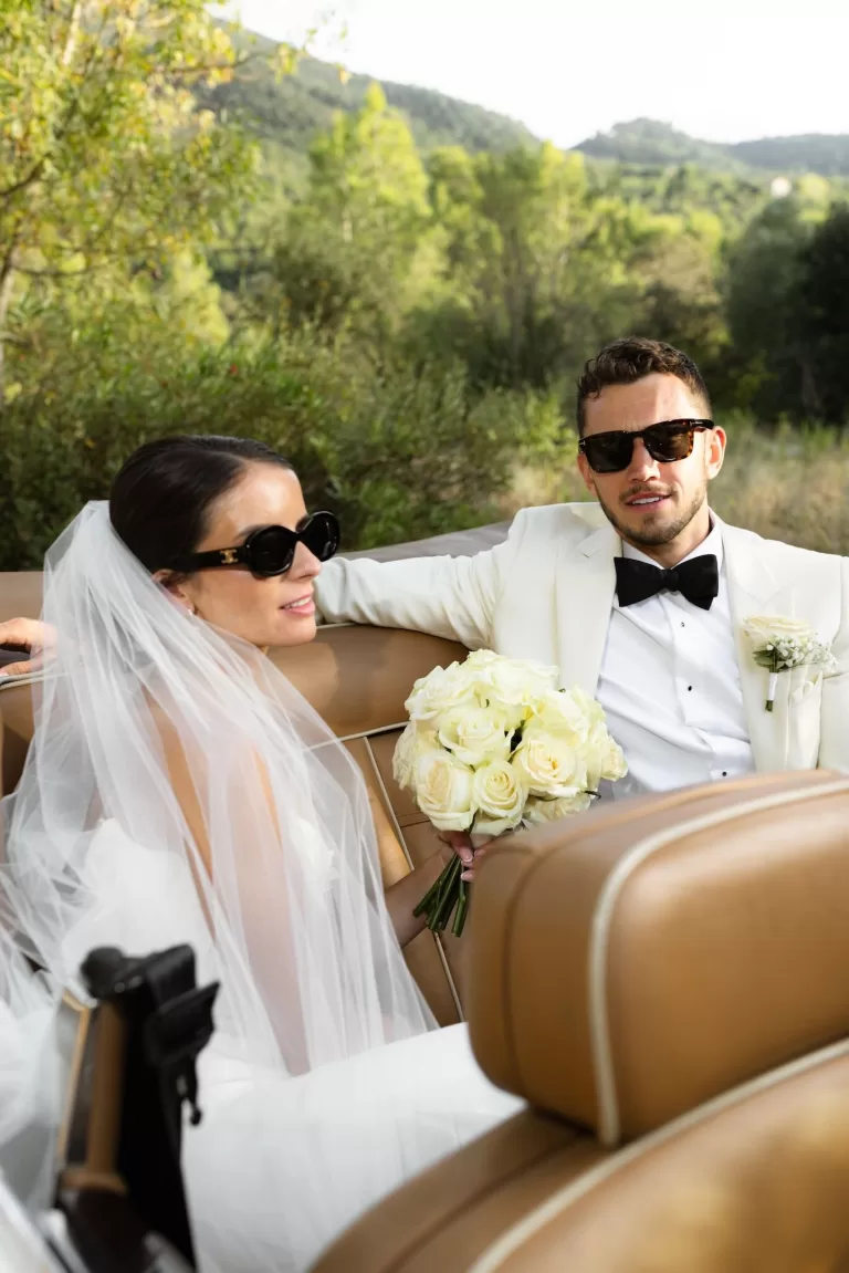 Bride and groom relaxing in a vintage convertible at Mas del Sord, holding a bouquet of white roses.